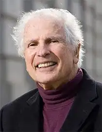 outdoor shot of a caucasian male with a full head of grey hair wearing a purple turtleneck sweater and jacket