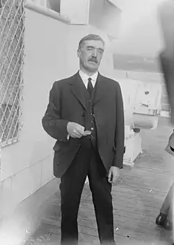 Three-quarter length photograph of a moustachioed man standing on the deck of a ship