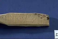 Image contains a piece of wood against a blue background. The piece of wood has several runes carved into its surface.