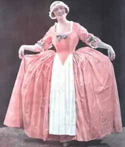 A white woman, standing, in a long pink gown with panniers and a cloth cap