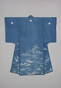 A medium blue figured silk kimono with a red lining viewed from the back. It has medium length sleeves and three crests, one in the centre back and two on each shoulder. The kimono is decorated on the waist down with designs of water, riverbanks, hills and trees in white, with small houses depicted in beige and brown.