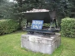 A monument commemorating the beginning of the oil shale industry, consisting of a grey concrete block about 1.5 metres wide and three-quarters of a metre deep. The block is topped by a black metal hopper filled with rocks. A blue plaque with white Estonian-language lettering just above the block explains its significance.