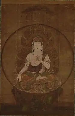 Frontal view of a deity seated on a pedestal in lotus position embellished with ornaments. The right arm is hanging down supported by the legs with the palm of the right hand facing forward. The left hand is placed in front of the body.