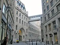 A narrow road with tall buildings of grey stone on both sides. The building on the left has a large entrance archway.