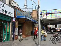 A brown-bricked building bordered in red bricks with a blue, rectangular sign reading "LADBROKE GROVE STATION" in white letters all under a blue sky