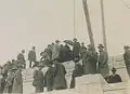 An old photo of a lot of men in suits and trenchcoats standing on top of the stone structure of the Cardston Alberta temple. Poles and wires are seen from the top.
