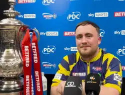 Luke Littler sitting in front of a panel of logos with microphones below his face and the Phil Taylor Trophy beside him