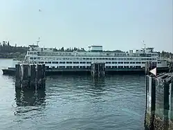 Side view of an older vessel with the standard Washington State Ferries livery, docked, image taken from another ferry. Worn-out paint, obvious rust, and an empty interior make it clear this ferry has been long retired.