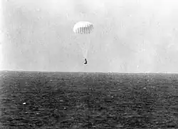 A conical black spacecraft falling towards the surface of the ocean under a single white parachute, seen from some distance away. Very little detail can be seen.