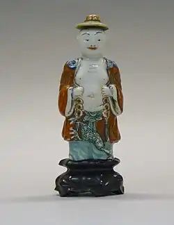 A porcelain snuff bottle in the shape of a man. He wears a stopper as a hat, and has richly painted robe, with a bare chest and belly.