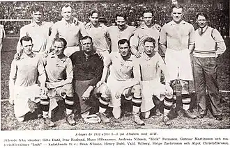 A black-and-white photograph of a football team lining up after a match. Six players wearing light shirts, white shorts and dark socks are standing behind a row of five kneeling players with the same apparel except for the man in the middle of the row of kneeling players who is wearing a dark shirt, dark shorts and dark socks. A man with casual clothing is standing to the right side of the row of standing players.