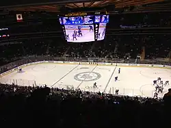 An arena set up for ice hockey. Spectators surround the rink, and players in blue and white jerseys can be seen on the ice.