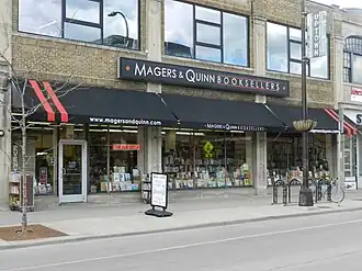 Photograph of the front of Magers & Quinn Booksellers, viewed from across the street. A black awning has two vertical red stripes on either side. White text reads www.magersandquinn.com. The shop has large glass windows, and a plethora of books on display. A neon sign reads, "We Buy Books". There are also carts of books for perusal on the sidewalk.