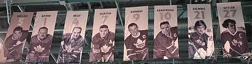 We are looking up at the ceiling of an arena where there are nine banners hanging. On each banner is the picture of a hockey player in a Toronto Maple Leaf uniform. Above each player's photo is a number and the player's name