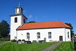 A white church with a red-tiled roof and a bell tower, set against a clear blue sky with green grass and a pathway in the foreground.