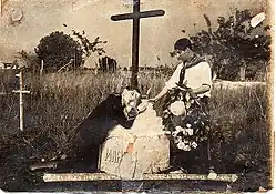 Black and white photo showing a man kneeling on a grave decorated with a cross, with another man standing next to him, holding a wreath of flowers.