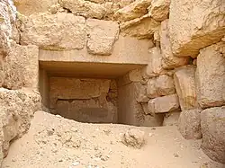 entrance of a stone lined corridor filed with desert sand amidst stone rubble