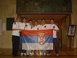 Six boys, standing on a line, all wearing white tops with red logos on their chest. They are holding a red, blue and white striped flag, which features a prominent crown and coat of arms.