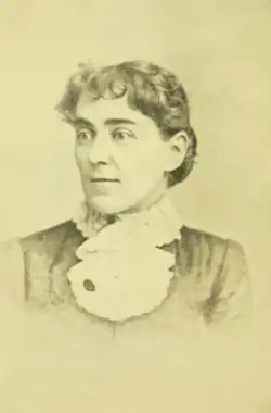 A young white woman with a curled fringe of dark hair, and dark eyebrows, she is wearing a high-collared garment with a kerchief ruffle at her throat