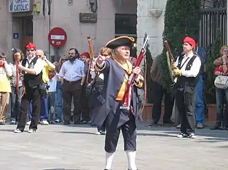 Photo shows a man in early 1800s garb with a short rifle. He wears a blue coat and trousers with a yellow waistcoat, a black tricorne hat with yellow trim, and white knee stockings.