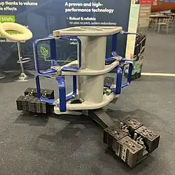 Photo of the HydroQuest turbine model sitting on the floor. It has a grey vertical central spine, supported on a squat three leg foundation. The central spine has 3 pairs of horizontal arms protruding either side. Between these are mounted 4 three-bladed vertical-axis turbines, which somewhat resemble an egg beater