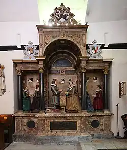 A large wall monument featuring a classical architectural framework with Corinthian columns supporting an arched canopy. The central scene depicts six kneeling figures in early 17th-century dress arranged in prayer within a colonnade. The central male figure, presumably Sir Roger Aston, wears dark armour and kneels facing a woman in an ochre-coloured gown, likely his wife. Four additional female figures, probably their daughters, kneel on either side wearing gowns in green, dark blue, red, and other colours, all with characteristic white ruffs of the period. Above the figures are heraldic shields displaying family coats of arms. The monument is constructed from polychrome marble with detailed carved decoration throughout the architectural framework. A black inscription tablet is mounted on the tomb chest below the kneeling figures. The overall composition demonstrates the classical style characteristic of the Cure workshop's funerary monuments of this period.