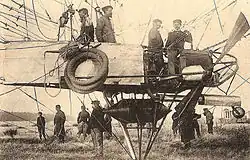 An airship gondola is shown in close-up from mid-left to mid-right; several items of equipment hang from the gondola's side; below the gondola a pyramid of steel tubing protrudes, shielding a cylindrical fuel tank; four crew members are in the gondola; above it mooring ropes and a rip-line are visible. A flat-ended propeller is also visible on the right