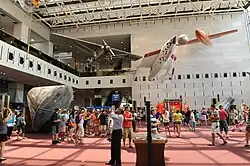 A crowd of people gather around various exhibits in a large museum hall. A grey single-engine monoplane, a white rocket-powered aircraft and an orange rocket-powered aircraft all hang from the ceiling above the crowd.