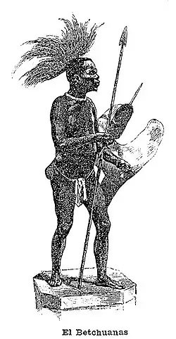 A black-and white print of a figure of a standing black man on a stand. He faces left. He wears a loincloth, a spear, a shield and feathers on his head.