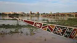 A colourful boat on the bank of a marshy river. The city skyline is behind the river.