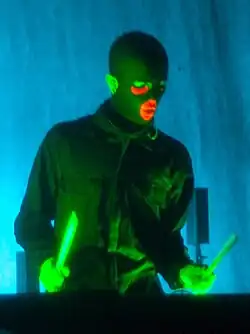 Olof Dreijer wearing a dark button-up, long-sleeved shirt and dark, ski-mask-like head covering, performing onstage while holding two drum sticks at a DJ station