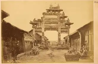 Ornamental gateway (pailou) from the Han dynasty (202 BCE – 220 CE) across a street lined with small shops. Hanzhong, Shaanxi Province, China (1875).