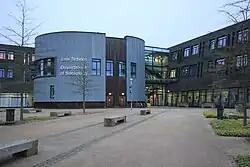 photo shows the front of the school of law and department of sociology building on campus east. The building is clad in wood with large portions of glass. There is a large plaza with semi-mature trees in the foreground.