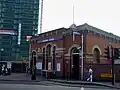 A red bricked cube shaped building with a rectangular, dark blue sign reading "PLAISTOW STATION" in white letters and people walking on the pavement in front