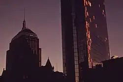 Two dark buildings rise into the early evening sky. The tower on the right, the Hancock Tower, has plywood on its side.