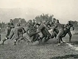 A cluster of American football players collide on a dirt field