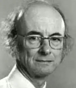 Black and white studio portrait of an unsmiling, clean-shaven, elderly, bespectacled man, with receding, unkempt, grey hair and striking, black eyebrows.