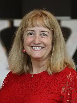 Rosie Stephenson-Goodknight in front of the Cardboard W in Wikimania 2023. She is a middle-aged white woman with blonde hair wearing a red dress.