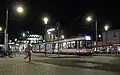 A picture of a tram at night. The Freiburg University library is visible in the distance. In the foreground, a blurry person walking is visible. They walk on cobblestone. Streetlights are ablaze, and the tram also lights up the scene.