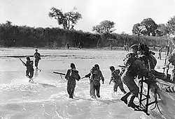 men climbing down side of small boat into the surf on to a sandy beech with trees in the background