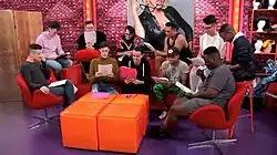 Screenshot of various contestants out of drag standing in a set, with an impartial image of RuPaul in the background.