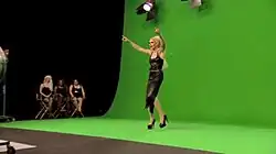 Screenshot of a drag performer standing in front of a green screen, with three other seated drag performers watching from the side