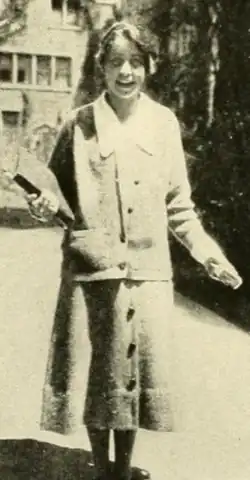 A smiling young white woman photographed outdoors, wearing a cardigan suit over a white blouse with a long pointed collar; she holds books with one arm