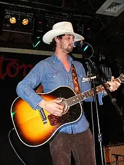 A man wearing a blue collared shirt and a cowboy hat is playing a guitar and singing into a microphone.
