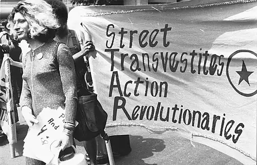Sylvia Rivera in 1970 at S.T.A.R. Rally, standing beside a banner that reads "Street Transvestite Action Revolutionaries