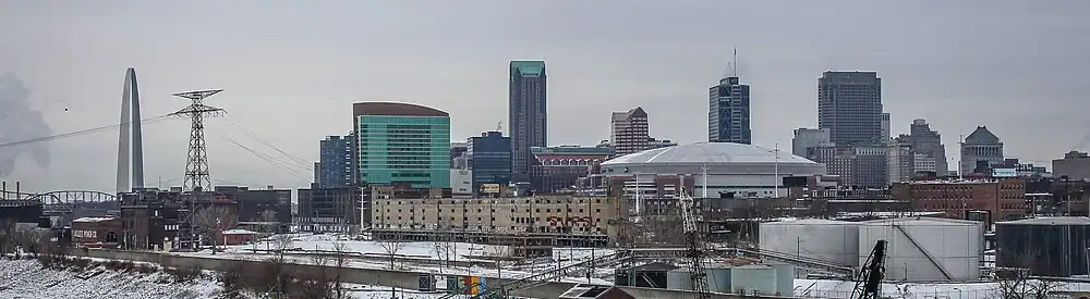 A large arch is in the center, across from a river. A clump of tall buildings is scattered behind it.