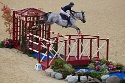A gray horse, ridden by a woman, in mid-air over a red and white striped fence.