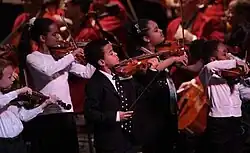 five children dressed in black and white play violins standing in front of seated adults in orange jackets