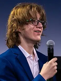 Portrait of Gibson facing slightly away from the camera to the right: wearing a blue jacket and glasses, while smiling and holding a microphone.