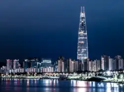 A nighttime shot of the Lotte tower from across the river. The tower is lit up and is far taller than any of the apartment buildings around it. By the riverside, a row of streetlights are shining on the river.